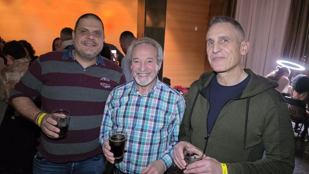 Tres hombres sonrientes sosteniendo bebidas en una fiesta temática de los 80 y 90.