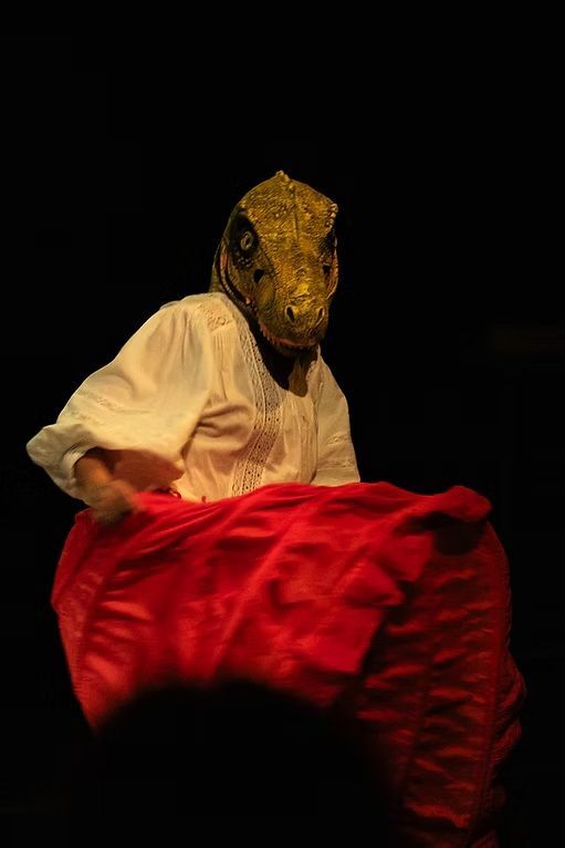 Actriz con máscara de dinosaurio y vestido rojo en escena teatral