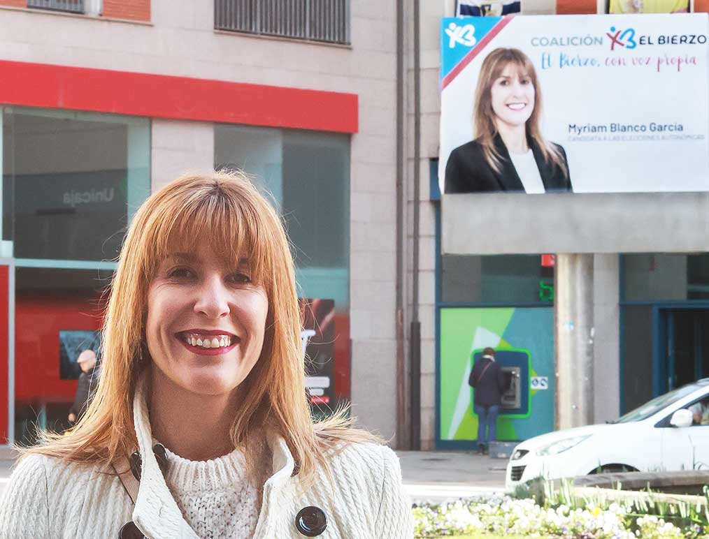 Mujer sonriendo frente a un cartel de campaña de Coalición por El Bierzo.
