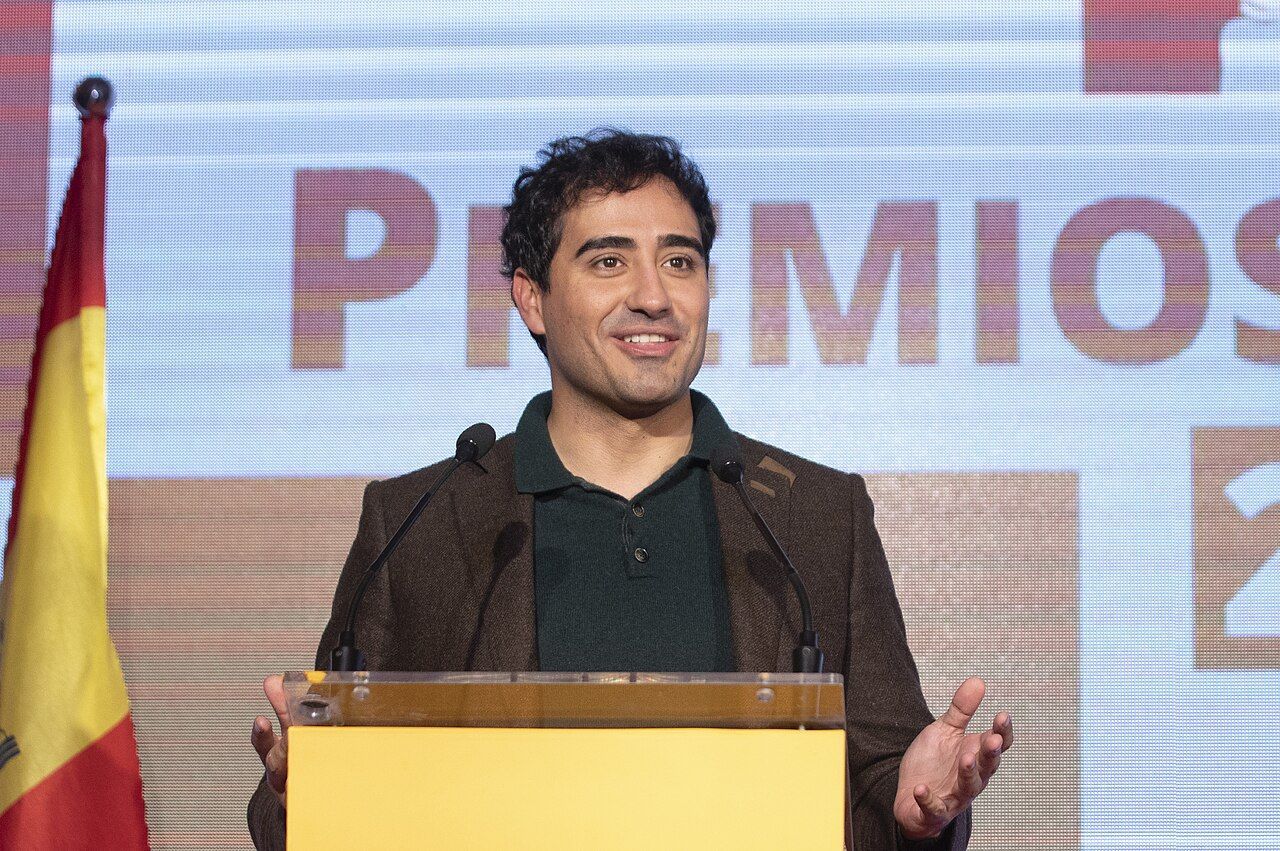Hombre sonriente en un evento con micrófono y bandera de España