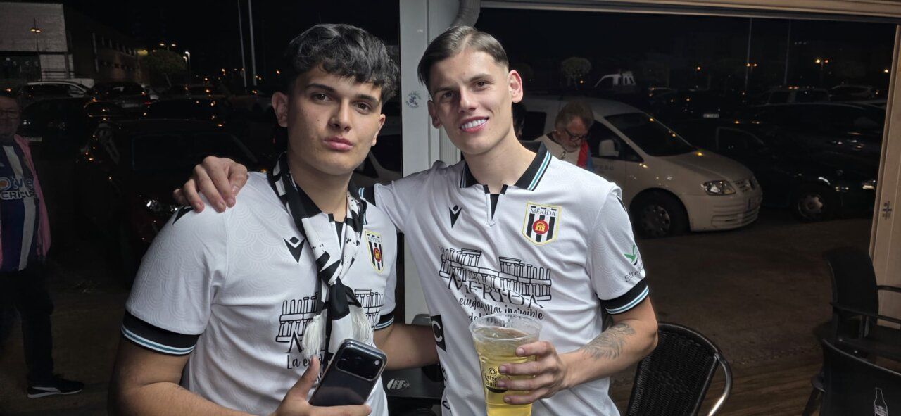 Dos jóvenes posando con camisetas del Mérida y una bebida en la mano.
