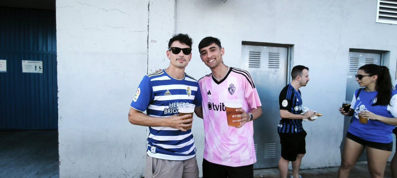 Dos jóvenes sosteniendo cervezas en un bar, uno con camiseta azul y otro con camiseta rosa.