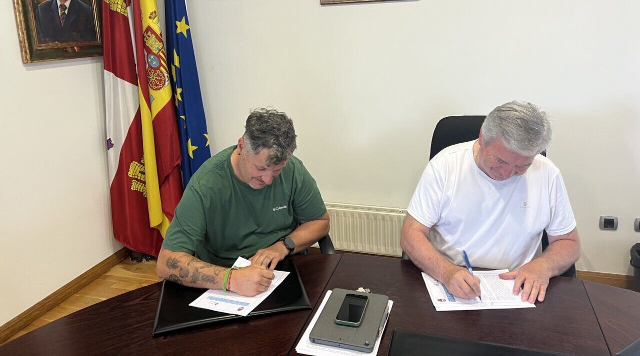 Dos personas firmando un convenio en una oficina con banderas