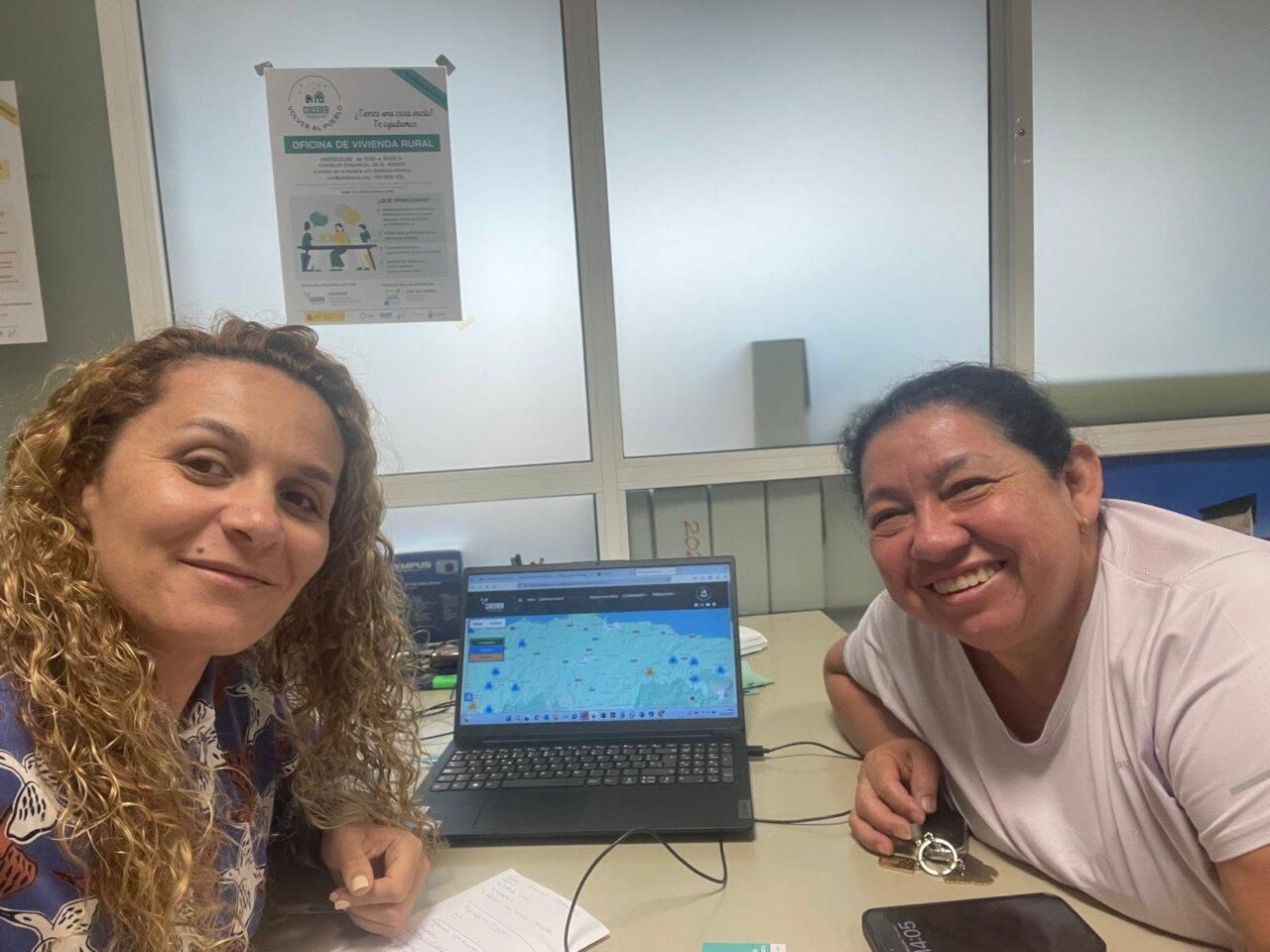 Dos mujeres sonrientes en una oficina con un ordenador y documentos