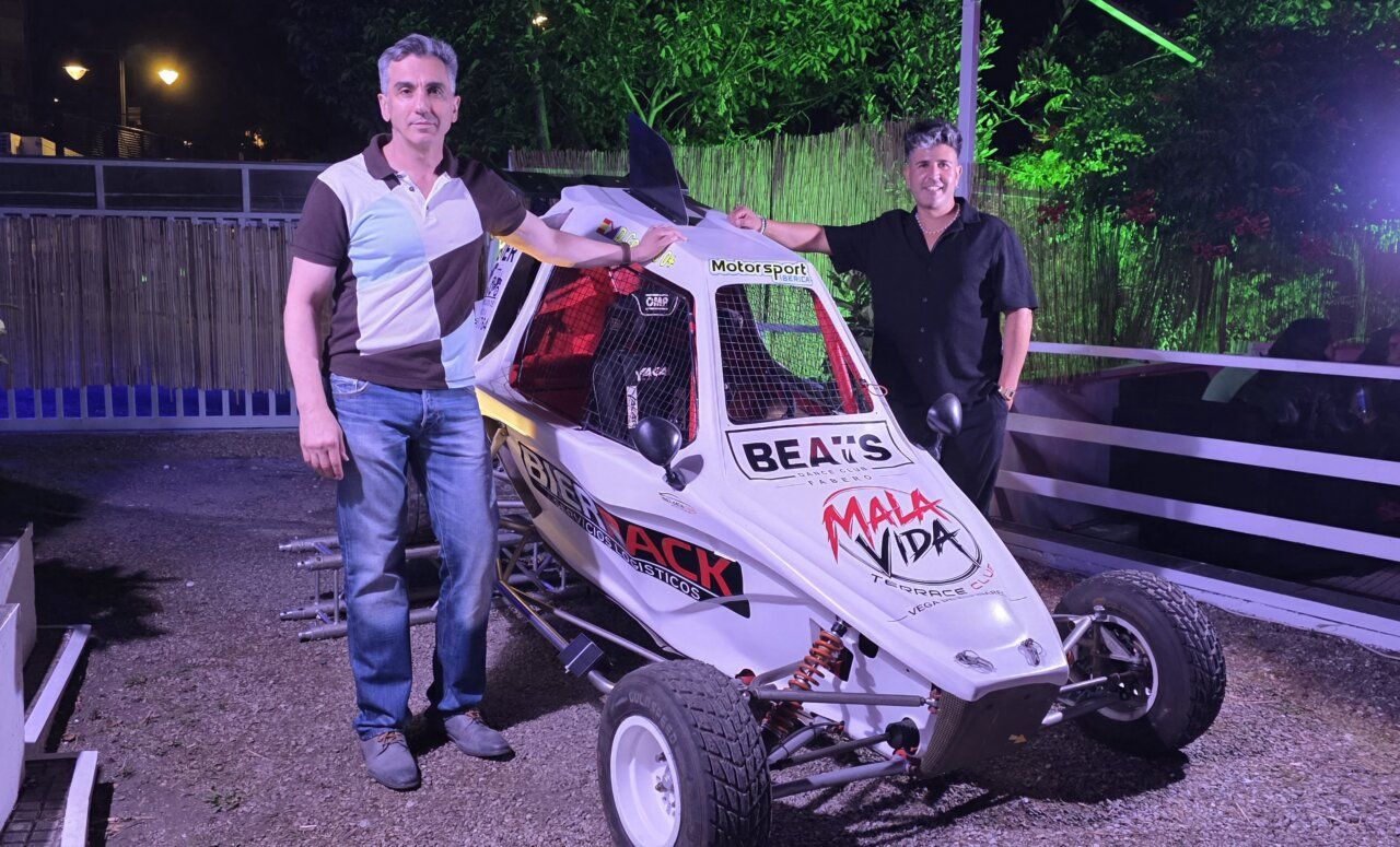 Dos hombres junto a un vehículo de competición en un evento nocturno.
