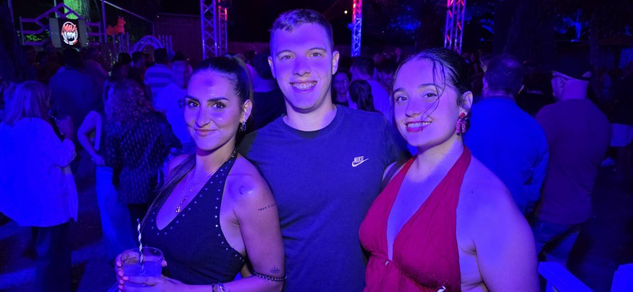 Tres personas sonriendo en un evento nocturno con luces de colores