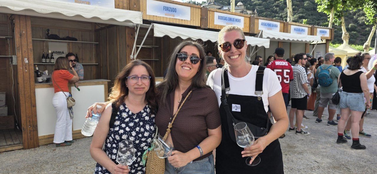 Tres mujeres sonrientes en un evento al aire libre con stands