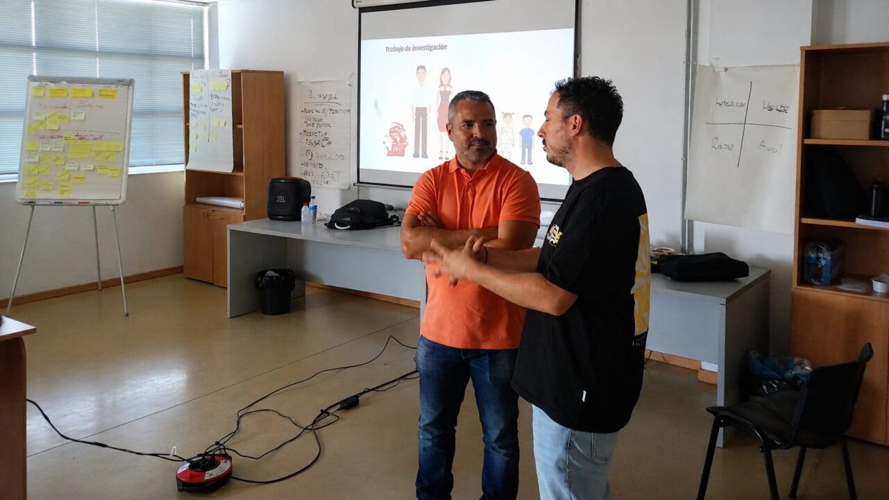 Dos hombres conversando en una sala de reuniones con presentaciones en la pantalla.