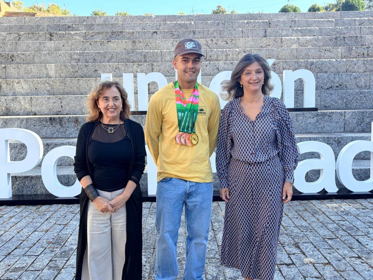 Joven con varias medallas posa entre dos mujeres ante unas letras blancas.