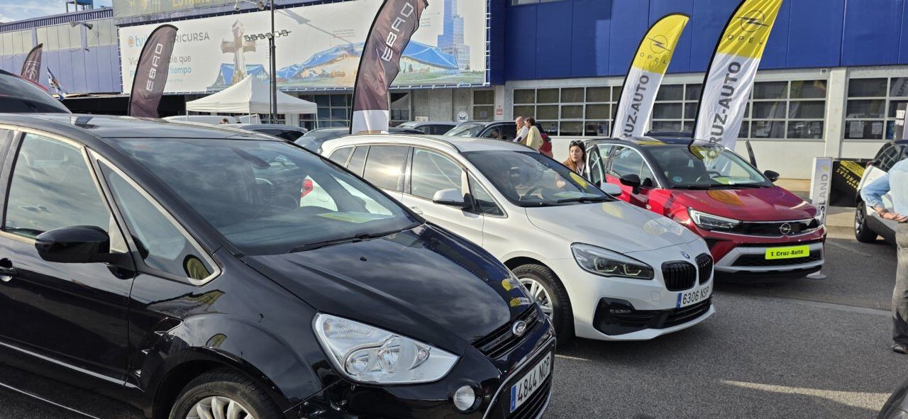 Varios coches expuestos frente a un edificio, con banderas de Cruz Auto.