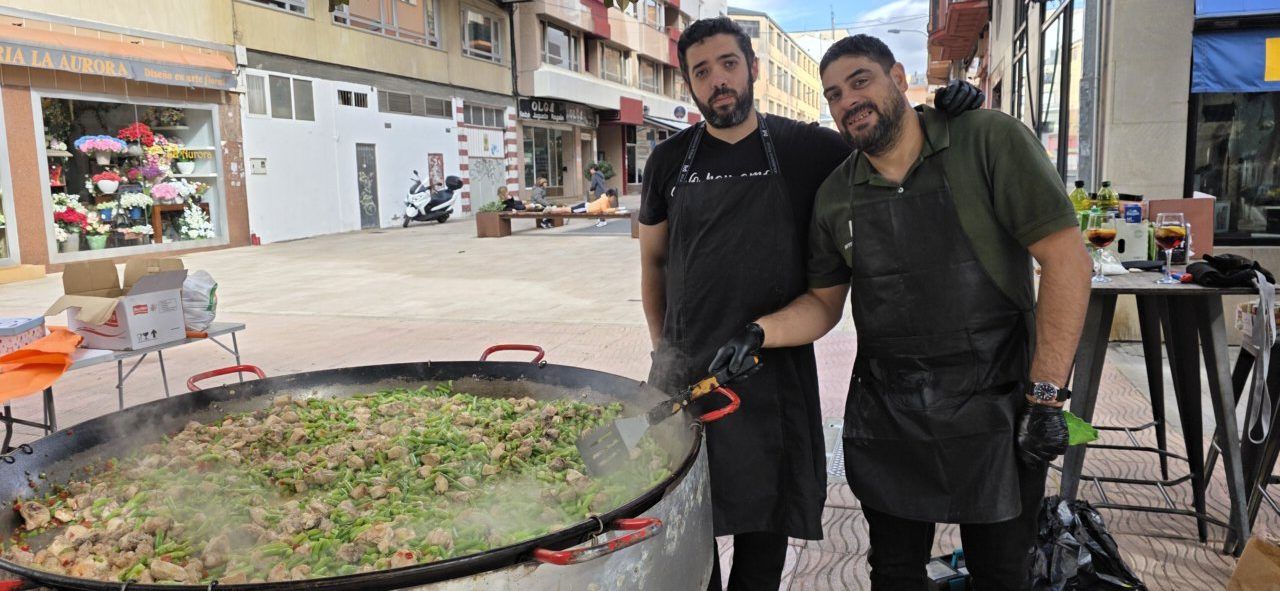 Dos hombres con delantales posan junto a una gran paellera con arroz y verduras