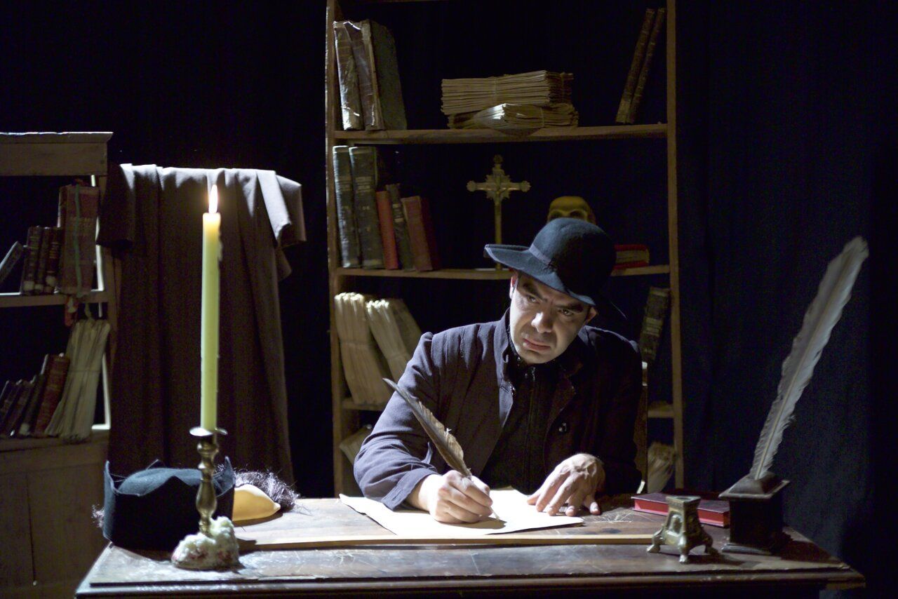 Actor sentado escribe con pluma en un escritorio rodeado de libros y una vela.