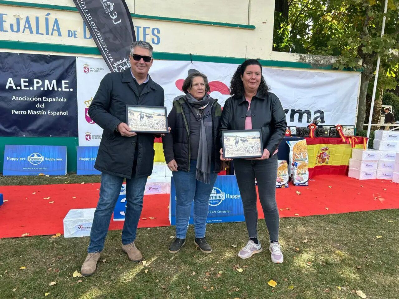 Tres personas posan con cuadros frente a carteles de una exposición canina.