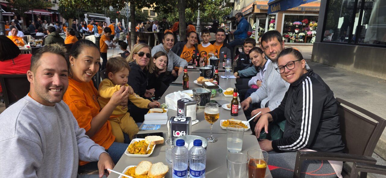 Varias personas comparten una comida en mesas al aire libre, algunas con camisetas naranjas.