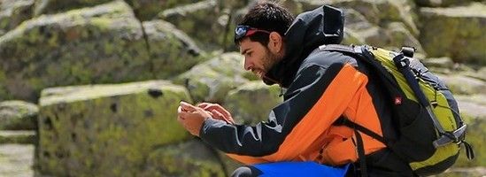 Hombre con chaqueta naranja y mochila consulta un teléfono en un entorno rocoso.