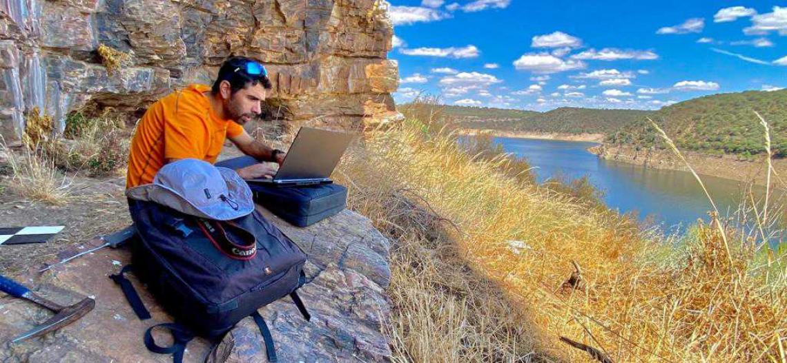 Un hombre trabaja con un portátil sentado sobre rocas junto a una masa de agua.