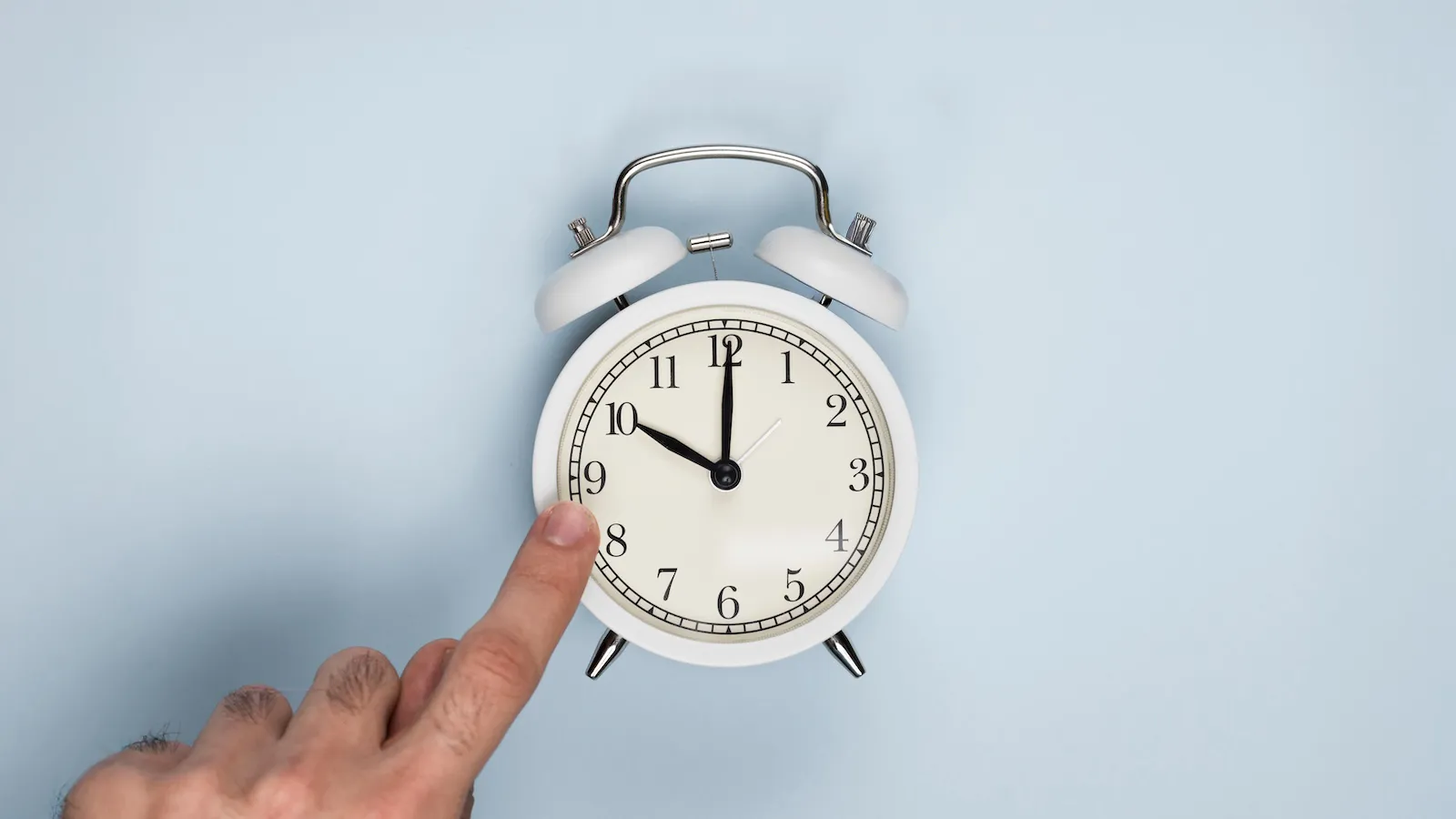 Una mano señala las diez en un reloj despertador blanco sobre fondo azul.
