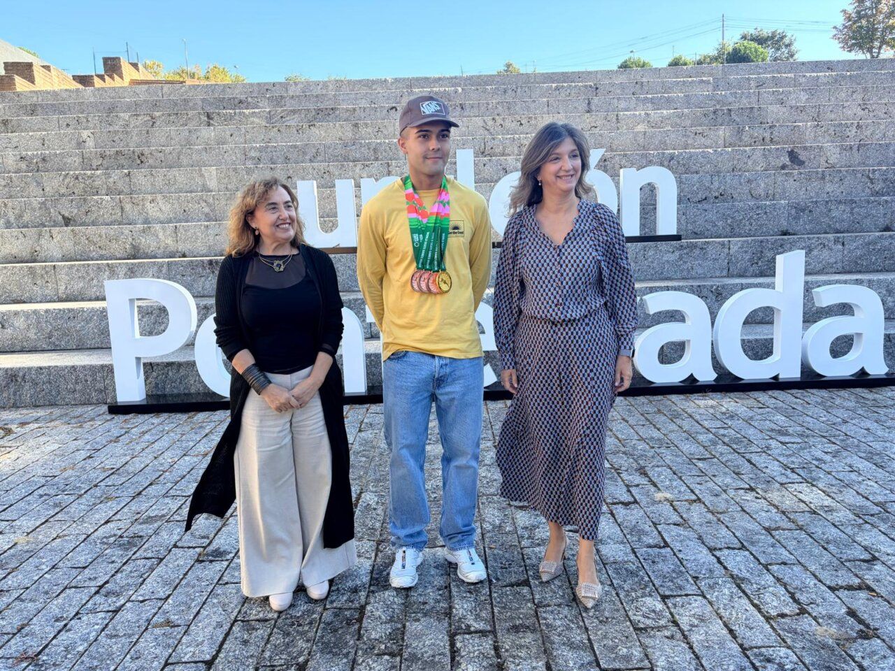 Tres personas posan ante unas gradas; el hombre del centro lleva varias medallas.