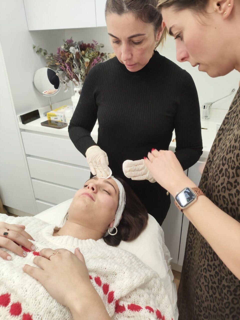 Una mujer con guantes toca la frente de una mujer recostada durante un cuidado facial.