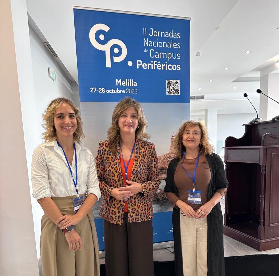 Tres mujeres posan frente a un cartel de las Jornadas Nacionales de Campus Periféricos en Melilla.