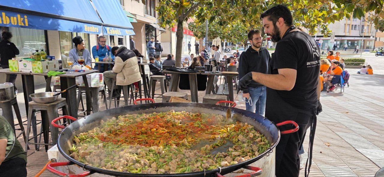 Una gran paella se cocina al aire libre junto a varias personas reunidas en una terraza.