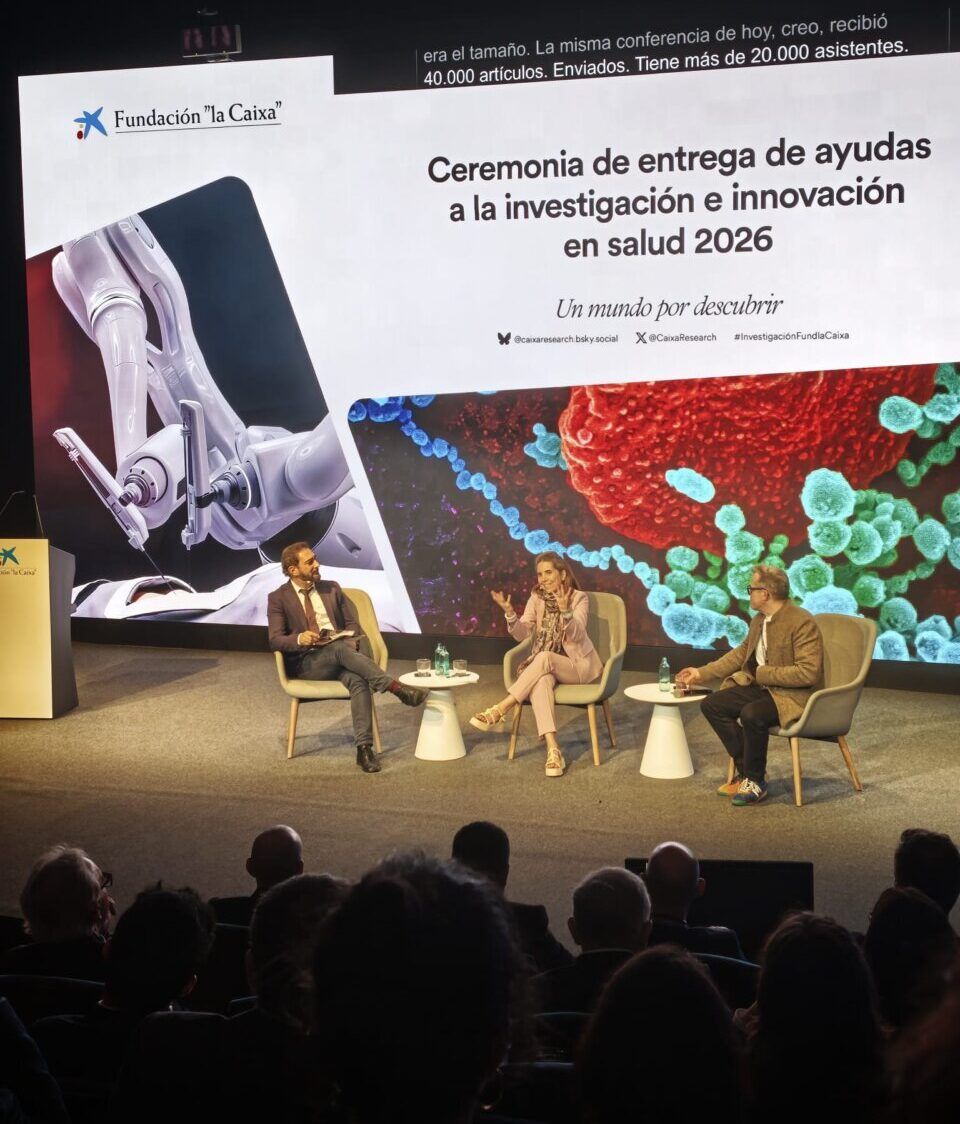 Tres personas conversan sentadas en un escenario ante una pantalla de la Fundación la Caixa.