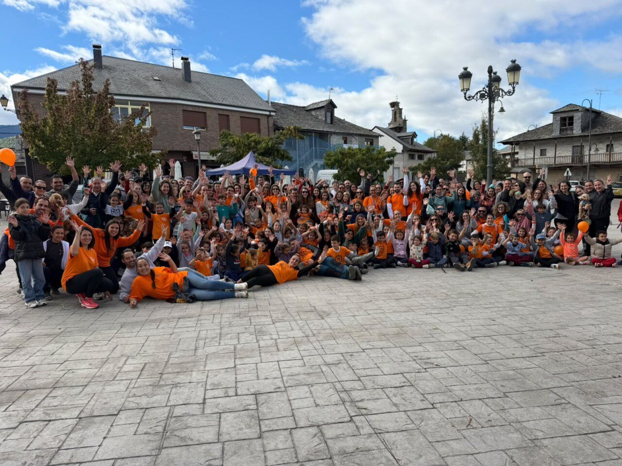 Grupo numeroso posa en una plaza y saluda a la cámara, muchos con camisetas naranjas.