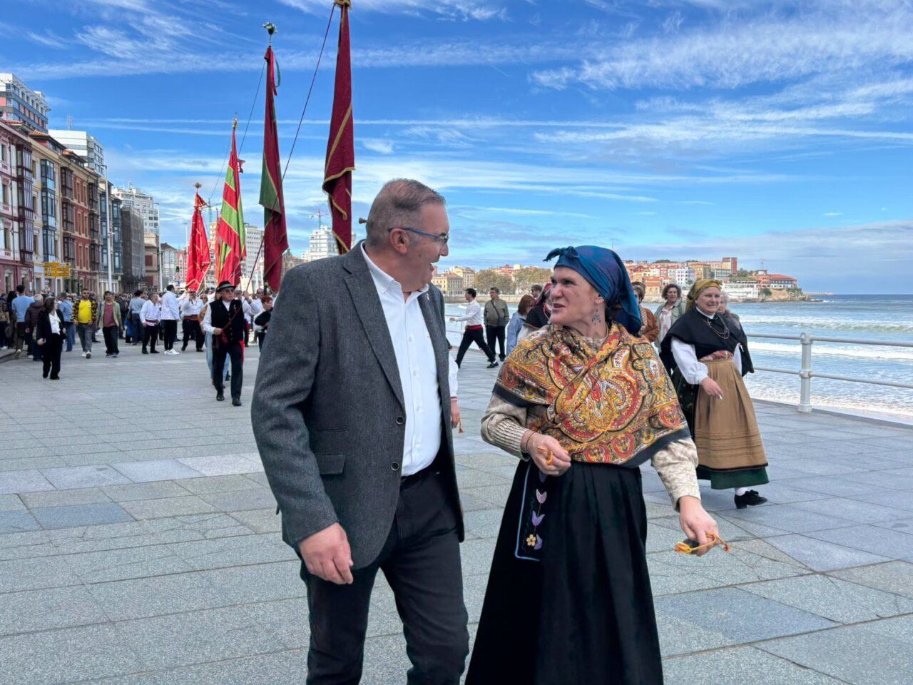 Personas junto a pendones y vestidas con trajes tradicionales en un paseo marítimo.
