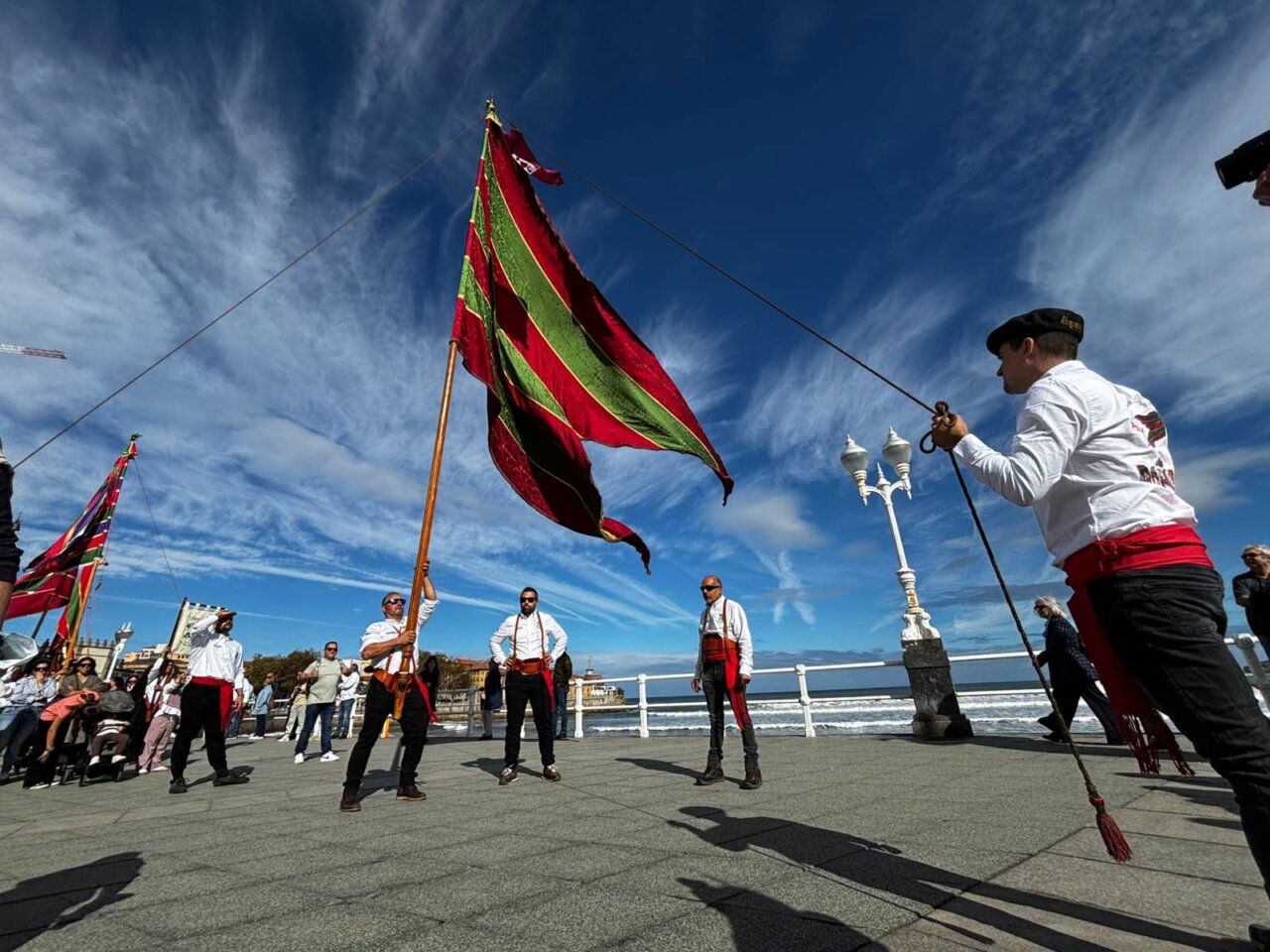 Personas sostienen grandes pendones rojos y verdes en un paseo junto al mar.