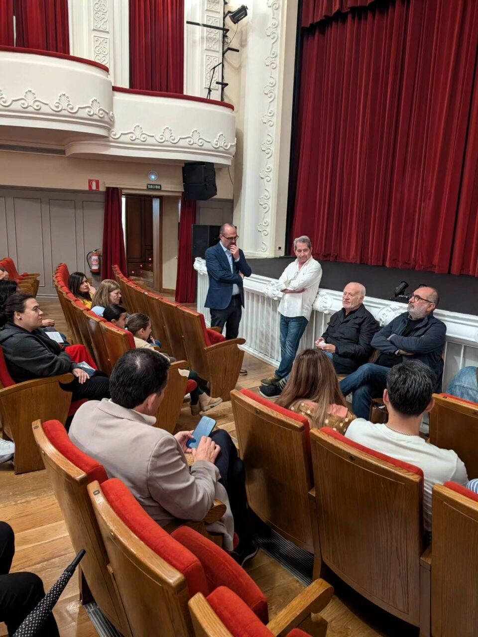 Personas sentadas en un teatro mientras varios hombres conversan junto al escenario.