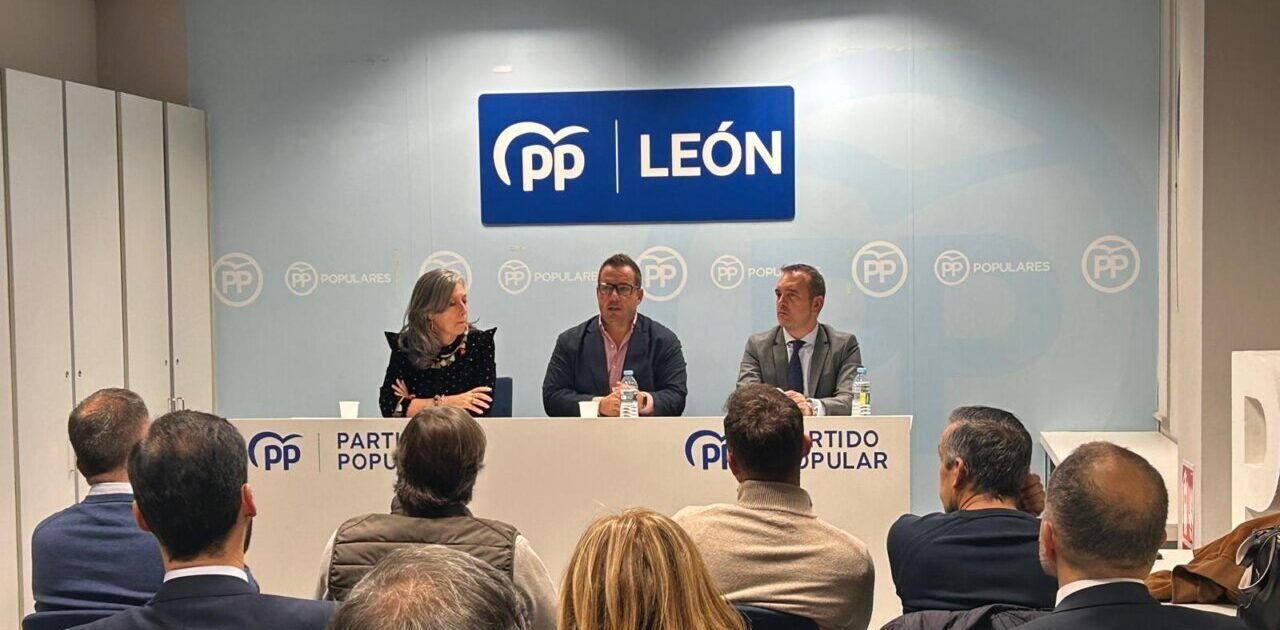 Tres personas sentadas ante una mesa con asistentes y rótulos del PP en León