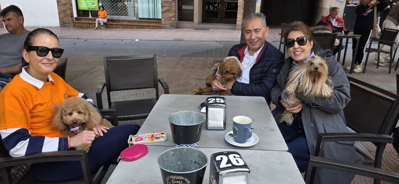 Varias personas sentadas en una terraza acompañadas por perros, con mesas y tazas delante.