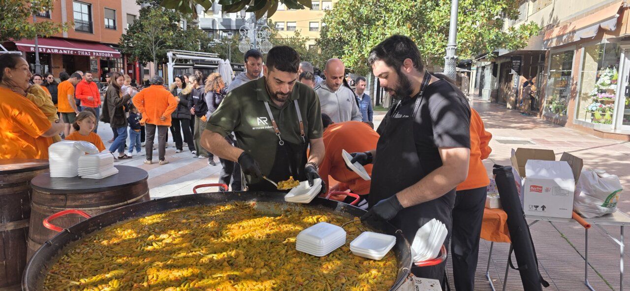 Dos hombres sirven paella en una gran sartén mientras varias personas esperan en una plaza.