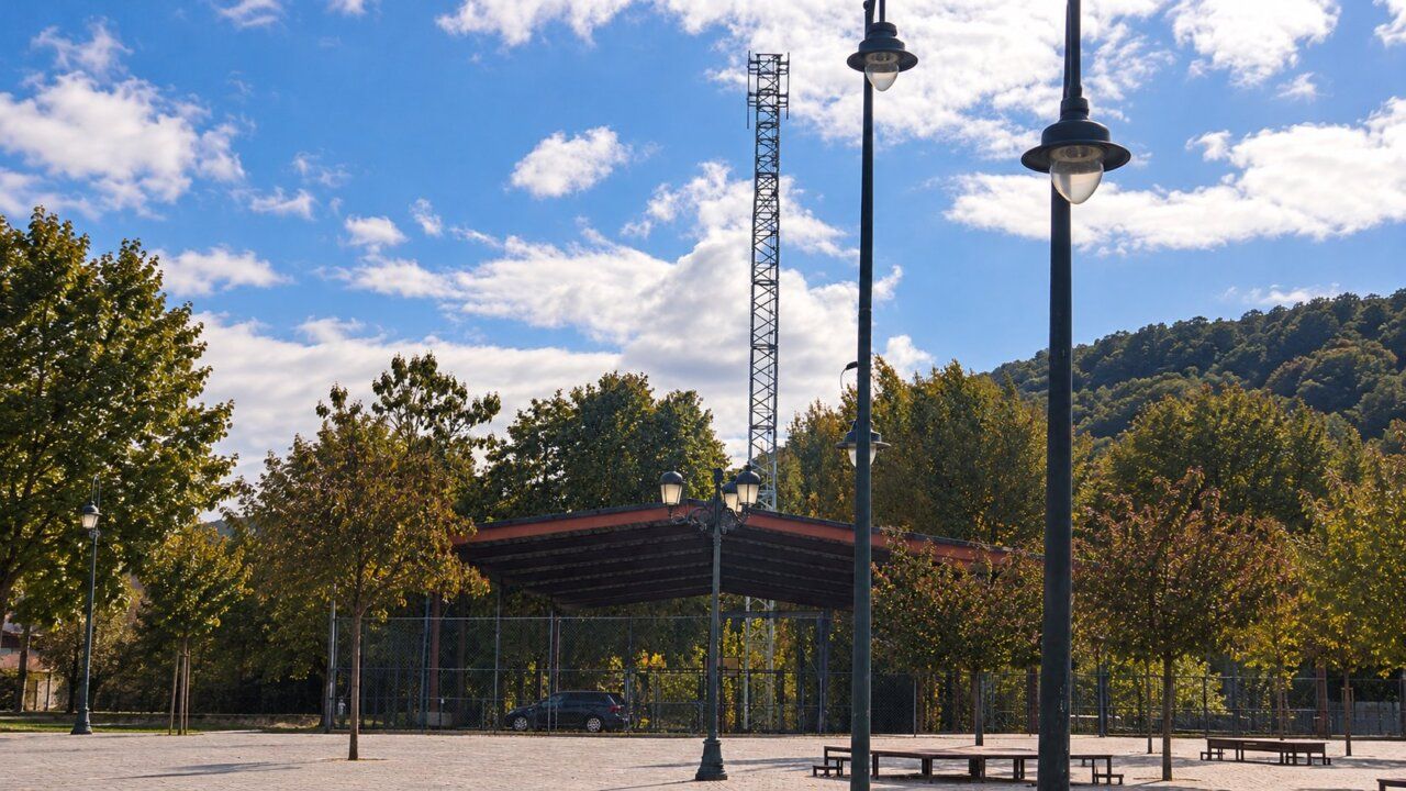 Plaza arbolada con farolas, bancos y una torre metálica al fondo