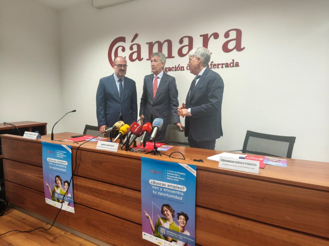 Tres hombres con traje ante micrófonos y carteles de una feria de empleo