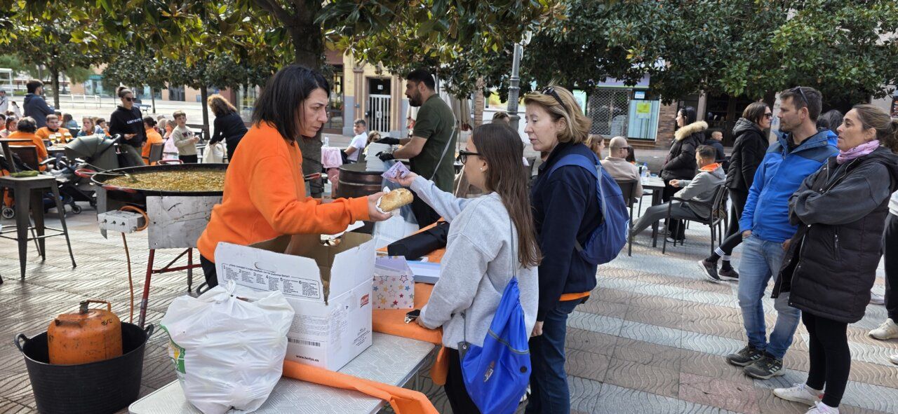 Una mujer entrega pan a una joven junto a una paella y varias mesas.