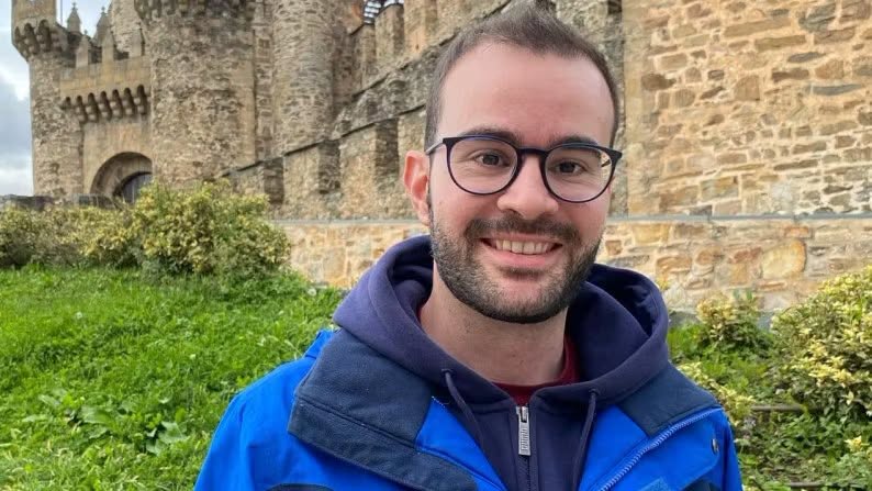 Hombre con gafas y chaqueta azul posa frente a un edificio de piedra.