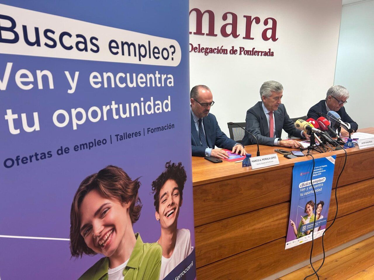 Tres hombres participan en una rueda de prensa junto a un cartel sobre ofertas de empleo.