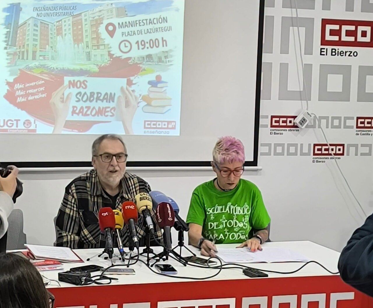 Dos personas ante micrófonos durante una rueda de prensa con un cartel proyectado.