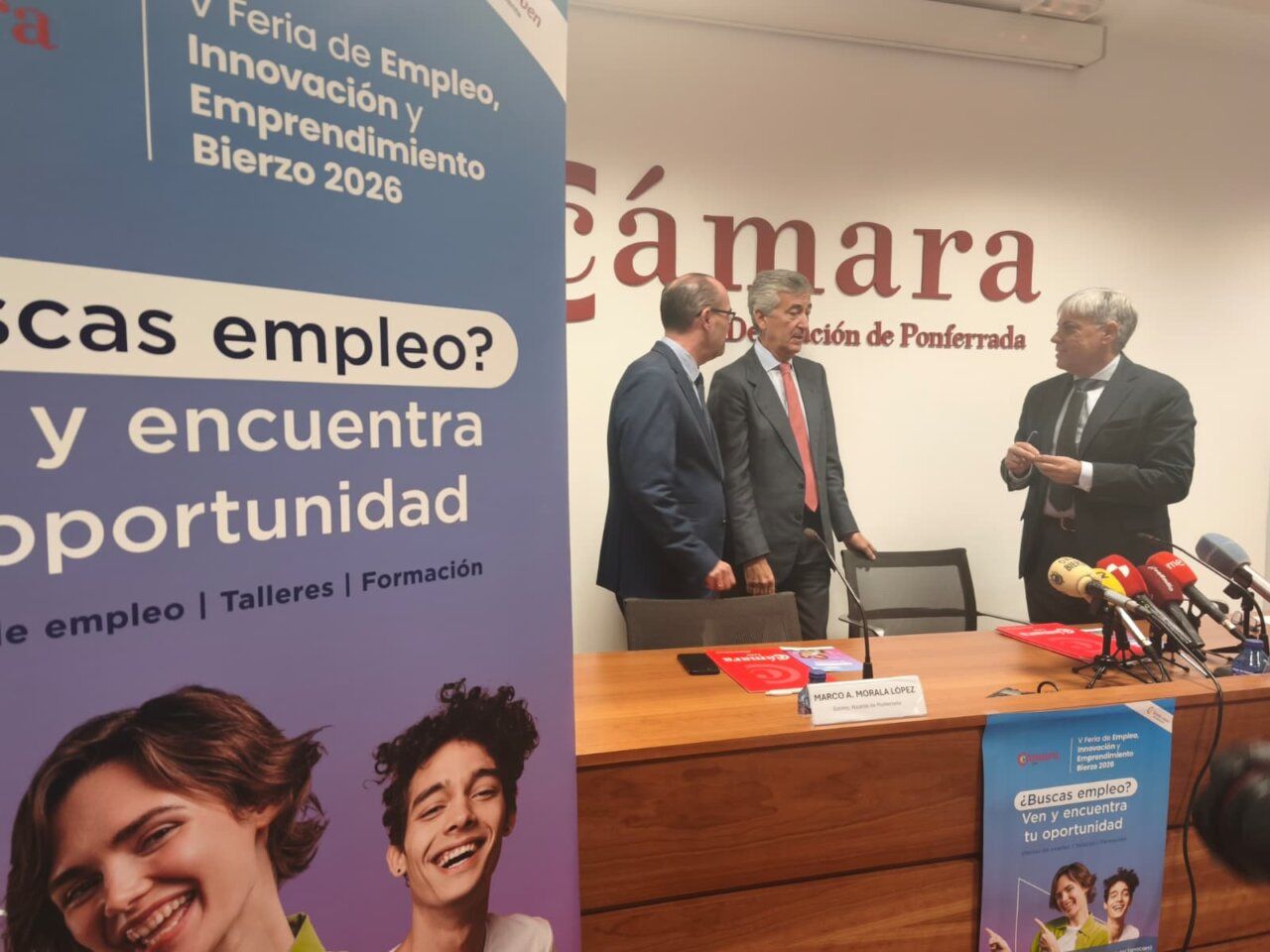 Tres hombres junto a una mesa con micrófonos y un cartel de la Feria de Empleo