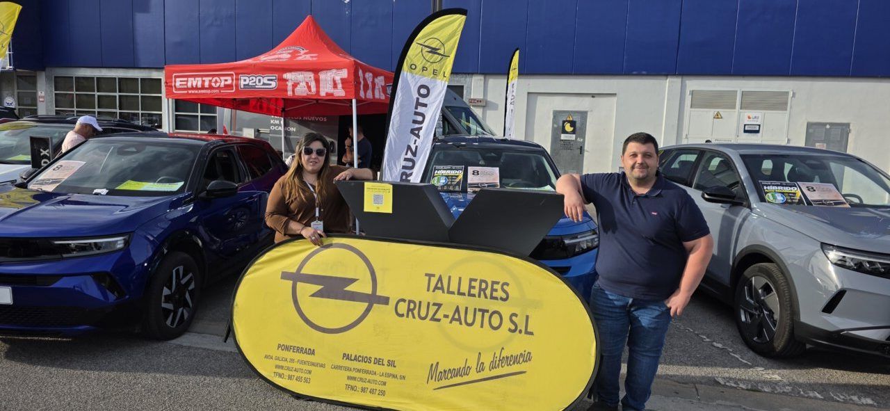 Dos personas junto a un cartel de Talleres Cruz-Auto, rodeadas de varios coches.