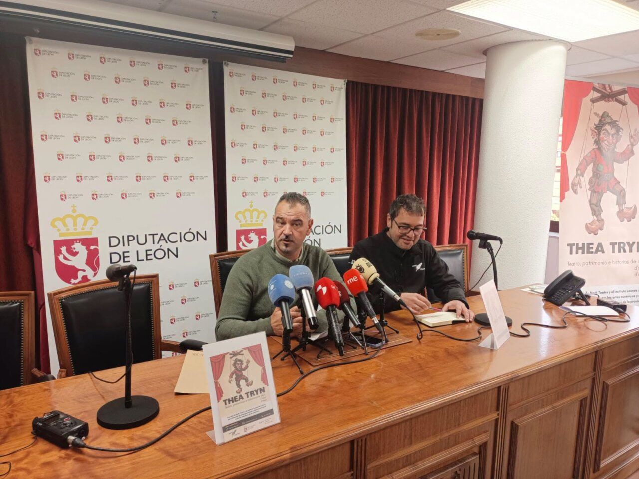 Dos hombres sentados ante micrófonos junto a carteles de Thea Tryn y Diputación de León